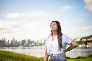 Shannon Gatta, a Hispanic woman with long, brown hair draped across her shoulder, stands at Gas Works Park with the Seattle skyline outlined behind her.